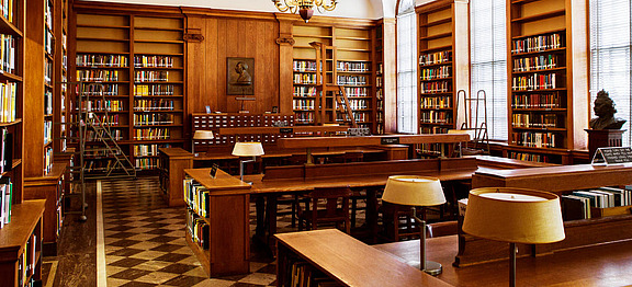Large library, brown wooden shelves filled with multicolored books, long brown wooden tables surrounded by chairs, tall glass windows